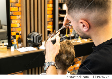 The picture of a small child with blue eyes and blond hair in a chair at the barbershop is in the process of cutting his hair with scissors. 114978776