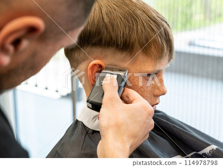 The picture of a blonde child with blue eyes and freckles is in a barbershop beauty salon. The miller holds a milling trimmer in his hand. The picture of a blonde child with blue eyes and freckles is in a barbershop beauty salon. The miller holds a milling trimmer in his hand. 114978798
