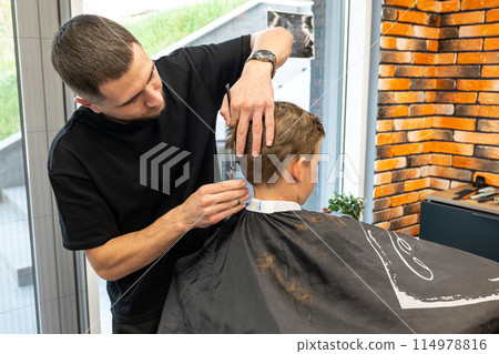 Young male barber making a haircut for cute little boy in the modern barbershop. Child in barbershop. Haircuts for Kids Young male barber making a haircut for cute little boy in the modern barbershop. Child in barbershop. Haircuts for Kids 114978816