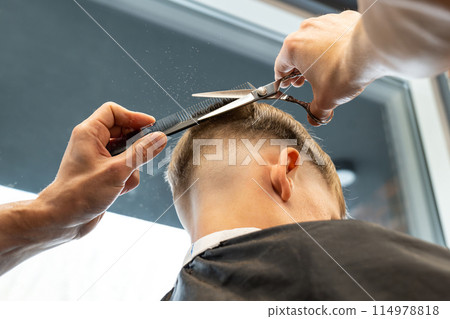 Stylist cuts male client hair using comb and scissors in barbershop closeup. Barber with professional tools serves man in salon 114978818