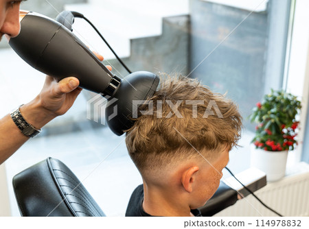 The picture of a professional barber is with a hair diffuser that he uses to make curly hair of a blond and freckled boy. The boy wants to have curly and beautiful hair. 114978832