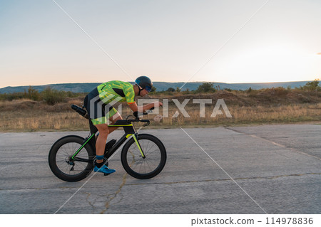 Triathlete riding his bicycle during sunset, preparing for a marathon. The warm colors of the sky provide a beautiful backdrop for his determined and focused effort. Triathlete riding his bicycle during sunset, preparing for a marathon. The warm colors of the sky provide a beautiful backdrop for his determined and focused effort. 114978836