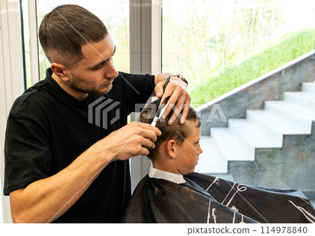 Man barber cutting little boy's hair using comb and scissors. Child getting haircut from adult male, likely barber. Professional hairdresser and cute client at modern barbershop. 114978840