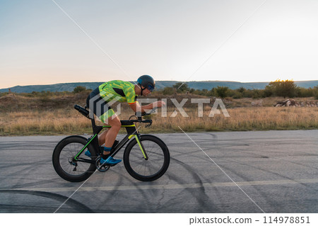 Triathlete riding his bicycle during sunset, preparing for a marathon. The warm colors of the sky provide a beautiful backdrop for his determined and focused effort. 114978851
