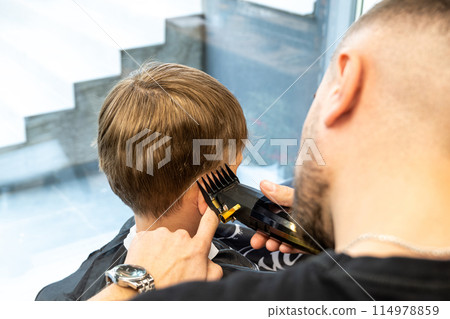 Attractive school boy is getting trendy haircut from mature hairdresser at fashionable hairdressing salon. 114978859