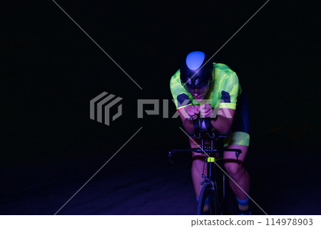 A triathlete rides his bike in the darkness of night, pushing himself to prepare for a marathon. The contrast between the darkness and the light of his bike creates a sense of drama and highlights the A triathlete rides his bike in the darkness of night, pushing himself to prepare for a marathon. The contrast between the darkness and the light of his bike creates a sense of drama and highlights the 114978903