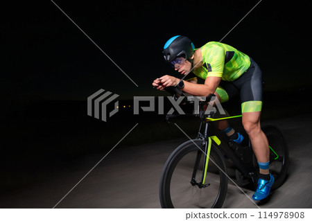 A triathlete rides his bike in the darkness of night, pushing himself to prepare for a marathon. The contrast between the darkness and the light of his bike creates a sense of drama and highlights the 114978908