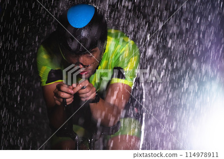 A triathlete braving the rain as he cycles through the night, preparing himself for the upcoming marathon. The blurred raindrops in the foreground and the dark, moody atmosphere in the background add 114978911
