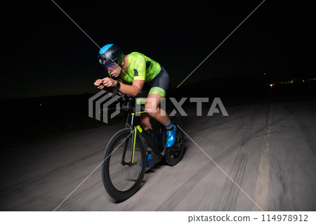 A triathlete rides his bike in the darkness of night, pushing himself to prepare for a marathon. The contrast between the darkness and the light of his bike creates a sense of drama and highlights the A triathlete rides his bike in the darkness of night, pushing himself to prepare for a marathon. The contrast between the darkness and the light of his bike creates a sense of drama and highlights the 114978912