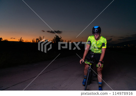 A triathlete rides his bike in the darkness of night, pushing himself to prepare for a marathon. The contrast between the darkness and the light of his bike creates a sense of drama and highlights the 114978916