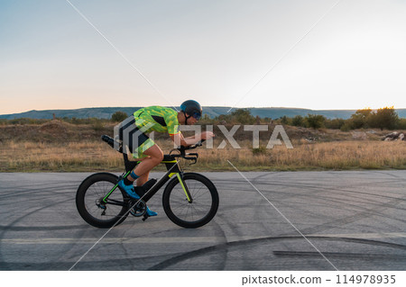 Triathlete riding his bicycle during sunset, preparing for a marathon. The warm colors of the sky provide a beautiful backdrop for his determined and focused effort. Triathlete riding his bicycle during sunset, preparing for a marathon. The warm colors of the sky provide a beautiful backdrop for his determined and focused effort. 114978935