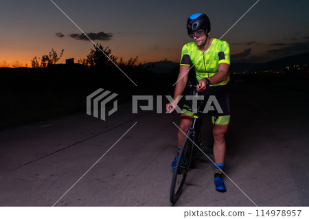 A triathlete rides his bike in the darkness of night, pushing himself to prepare for a marathon. The contrast between the darkness and the light of his bike creates a sense of drama and highlights the A triathlete rides his bike in the darkness of night, pushing himself to prepare for a marathon. The contrast between the darkness and the light of his bike creates a sense of drama and highlights the 114978957