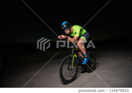 A triathlete rides his bike in the darkness of night, pushing himself to prepare for a marathon. The contrast between the darkness and the light of his bike creates a sense of drama and highlights the 114979020