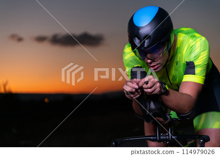 A triathlete rides his bike in the darkness of night, pushing himself to prepare for a marathon. The contrast between the darkness and the light of his bike creates a sense of drama and highlights the A triathlete rides his bike in the darkness of night, pushing himself to prepare for a marathon. The contrast between the darkness and the light of his bike creates a sense of drama and highlights the 114979026