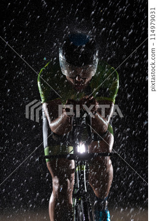 A triathlete braving the rain as he cycles through the night, preparing himself for the upcoming marathon. The blurred raindrops in the foreground and the dark, moody atmosphere in the background add 114979031