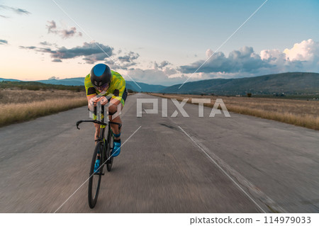 Triathlete riding his bicycle during sunset, preparing for a marathon. The warm colors of the sky provide a beautiful backdrop for his determined and focused effort. 114979033