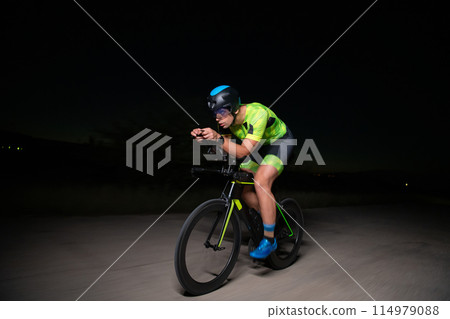 A triathlete rides his bike in the darkness of night, pushing himself to prepare for a marathon. The contrast between the darkness and the light of his bike creates a sense of drama and highlights the 114979088