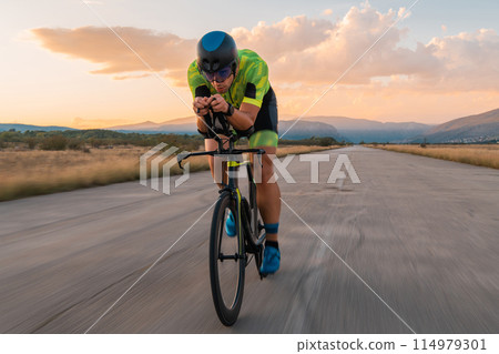 Triathlete riding his bicycle during sunset, preparing for a marathon. The warm colors of the sky provide a beautiful backdrop for his determined and focused effort. Triathlete riding his bicycle during sunset, preparing for a marathon. The warm colors of the sky provide a beautiful backdrop for his determined and focused effort. 114979301