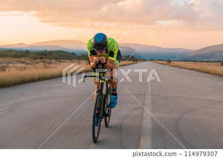 Triathlete riding his bicycle during sunset, preparing for a marathon. The warm colors of the sky provide a beautiful backdrop for his determined and focused effort. Triathlete riding his bicycle during sunset, preparing for a marathon. The warm colors of the sky provide a beautiful backdrop for his determined and focused effort. 114979307