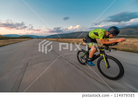 Triathlete riding his bicycle during sunset, preparing for a marathon. The warm colors of the sky provide a beautiful backdrop for his determined and focused effort. Triathlete riding his bicycle during sunset, preparing for a marathon. The warm colors of the sky provide a beautiful backdrop for his determined and focused effort. 114979385