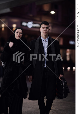 Happy multicultural business couple walking together outdoors in an urban city street at night near a jewelry shopping store window. Happy multicultural business couple walking together outdoors in an urban city street at night near a jewelry shopping store window. 114979402