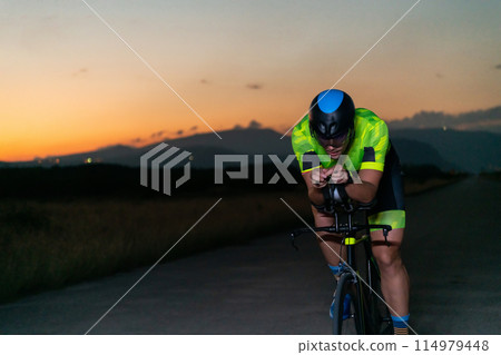 A triathlete rides his bike in the darkness of night, pushing himself to prepare for a marathon. The contrast between the darkness and the light of his bike creates a sense of drama and highlights the A triathlete rides his bike in the darkness of night, pushing himself to prepare for a marathon. The contrast between the darkness and the light of his bike creates a sense of drama and highlights the 114979448