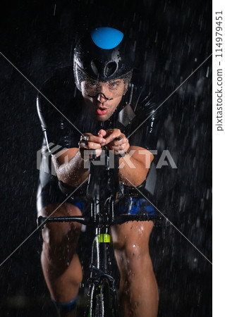 A triathlete braving the rain as he cycles through the night, preparing himself for the upcoming marathon. The blurred raindrops in the foreground and the dark, moody atmosphere in the background add A triathlete braving the rain as he cycles through the night, preparing himself for the upcoming marathon. The blurred raindrops in the foreground and the dark, moody atmosphere in the background add 114979451