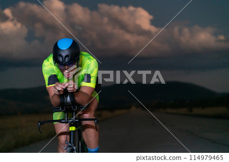 A triathlete rides his bike in the darkness of night, pushing himself to prepare for a marathon. The contrast between the darkness and the light of his bike creates a sense of drama and highlights the A triathlete rides his bike in the darkness of night, pushing himself to prepare for a marathon. The contrast between the darkness and the light of his bike creates a sense of drama and highlights the 114979465