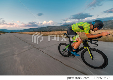 Triathlete riding his bicycle during sunset, preparing for a marathon. The warm colors of the sky provide a beautiful backdrop for his determined and focused effort. Triathlete riding his bicycle during sunset, preparing for a marathon. The warm colors of the sky provide a beautiful backdrop for his determined and focused effort. 114979472