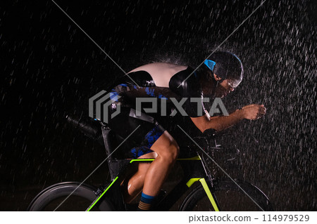 A triathlete braving the rain as he cycles through the night, preparing himself for the upcoming marathon. The blurred raindrops in the foreground and the dark, moody atmosphere in the background add A triathlete braving the rain as he cycles through the night, preparing himself for the upcoming marathon. The blurred raindrops in the foreground and the dark, moody atmosphere in the background add 114979529
