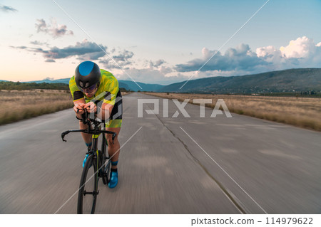 Triathlete riding his bicycle during sunset, preparing for a marathon. The warm colors of the sky provide a beautiful backdrop for his determined and focused effort. Triathlete riding his bicycle during sunset, preparing for a marathon. The warm colors of the sky provide a beautiful backdrop for his determined and focused effort. 114979622