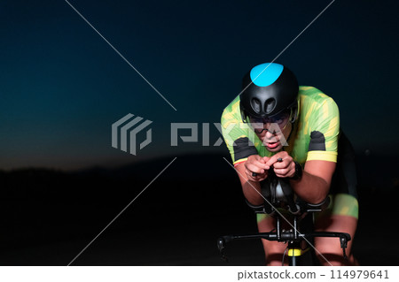 A triathlete rides his bike in the darkness of night, pushing himself to prepare for a marathon. The contrast between the darkness and the light of his bike creates a sense of drama and highlights the A triathlete rides his bike in the darkness of night, pushing himself to prepare for a marathon. The contrast between the darkness and the light of his bike creates a sense of drama and highlights the 114979641
