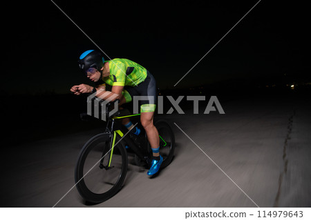 A triathlete rides his bike in the darkness of night, pushing himself to prepare for a marathon. The contrast between the darkness and the light of his bike creates a sense of drama and highlights the 114979643