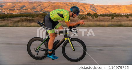 Triathlete riding his bicycle during sunset, preparing for a marathon. The warm colors of the sky provide a beautiful backdrop for his determined and focused effort. Triathlete riding his bicycle during sunset, preparing for a marathon. The warm colors of the sky provide a beautiful backdrop for his determined and focused effort. 114979735