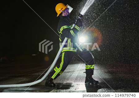 Firefighter using a water hose to eliminate a fire hazard. Team of firemen in the dangerous rescue mission. Firefighter using a water hose to eliminate a fire hazard. Team of firemen in the dangerous rescue mission. 114979740