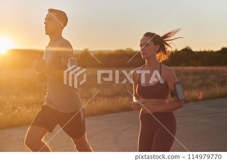 A handsome young couple running together during the early morning hours, with the mesmerizing sunrise casting a warm glow, symbolizing their shared love and vitality 114979770