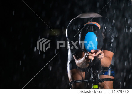 A triathlete braving the rain as he cycles through the night, preparing himself for the upcoming marathon. The blurred raindrops in the foreground and the dark, moody atmosphere in the background add A triathlete braving the rain as he cycles through the night, preparing himself for the upcoming marathon. The blurred raindrops in the foreground and the dark, moody atmosphere in the background add 114979780