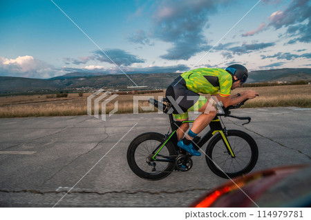 Triathlete riding his bicycle during sunset, preparing for a marathon. The warm colors of the sky provide a beautiful backdrop for his determined and focused effort. Triathlete riding his bicycle during sunset, preparing for a marathon. The warm colors of the sky provide a beautiful backdrop for his determined and focused effort. 114979781