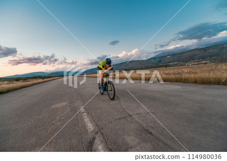 Triathlete riding his bicycle during sunset, preparing for a marathon. The warm colors of the sky provide a beautiful backdrop for his determined and focused effort. Triathlete riding his bicycle during sunset, preparing for a marathon. The warm colors of the sky provide a beautiful backdrop for his determined and focused effort. 114980036