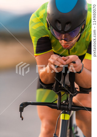 Close up photo of triathlete riding his bicycle during sunset, preparing for a marathon. The warm colors of the sky provide a beautiful backdrop for his determined and focused effort. Close up photo of triathlete riding his bicycle during sunset, preparing for a marathon. The warm colors of the sky provide a beautiful backdrop for his determined and focused effort. 114980070