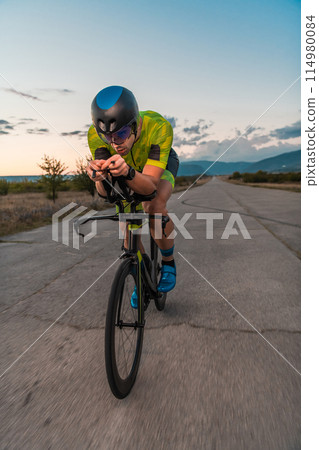 Triathlete riding his bicycle during sunset, preparing for a marathon. The warm colors of the sky provide a beautiful backdrop for his determined and focused effort. Triathlete riding his bicycle during sunset, preparing for a marathon. The warm colors of the sky provide a beautiful backdrop for his determined and focused effort. 114980084