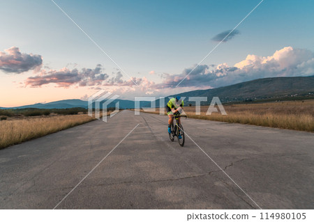 Triathlete riding his bicycle during sunset, preparing for a marathon. The warm colors of the sky provide a beautiful backdrop for his determined and focused effort. Triathlete riding his bicycle during sunset, preparing for a marathon. The warm colors of the sky provide a beautiful backdrop for his determined and focused effort. 114980105
