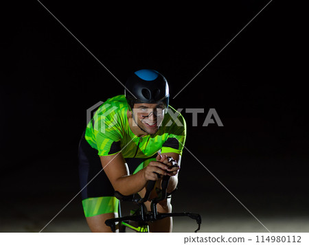 A triathlete rides his bike in the darkness of night, pushing himself to prepare for a marathon. The contrast between the darkness and the light of his bike creates a sense of drama and highlights the A triathlete rides his bike in the darkness of night, pushing himself to prepare for a marathon. The contrast between the darkness and the light of his bike creates a sense of drama and highlights the 114980112
