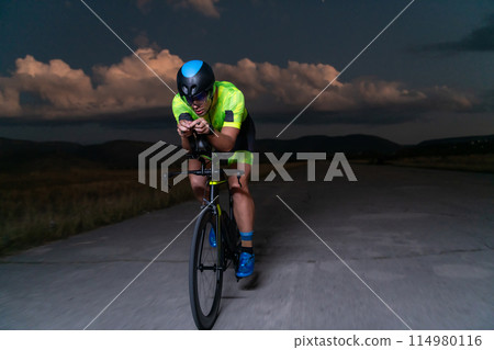 A triathlete rides his bike in the darkness of night, pushing himself to prepare for a marathon. The contrast between the darkness and the light of his bike creates a sense of drama and highlights the 114980116