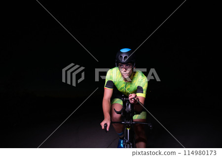 A triathlete rides his bike in the darkness of night, pushing himself to prepare for a marathon. The contrast between the darkness and the light of his bike creates a sense of drama and highlights the 114980173