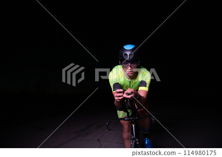 A triathlete rides his bike in the darkness of night, pushing himself to prepare for a marathon. The contrast between the darkness and the light of his bike creates a sense of drama and highlights the 114980175
