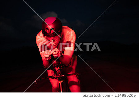 A triathlete rides his bike in the darkness of night, pushing himself to prepare for a marathon. The contrast between the darkness and the light of his bike creates a sense of drama and highlights the 114980209