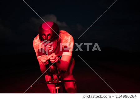A triathlete rides his bike in the darkness of night, pushing himself to prepare for a marathon. The contrast between the darkness and the light of his bike creates a sense of drama and highlights the 114980264