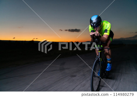 A triathlete rides his bike in the darkness of night, pushing himself to prepare for a marathon. The contrast between the darkness and the light of his bike creates a sense of drama and highlights the A triathlete rides his bike in the darkness of night, pushing himself to prepare for a marathon. The contrast between the darkness and the light of his bike creates a sense of drama and highlights the 114980279