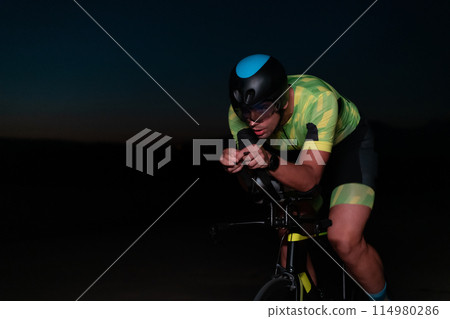 A triathlete rides his bike in the darkness of night, pushing himself to prepare for a marathon. The contrast between the darkness and the light of his bike creates a sense of drama and highlights the A triathlete rides his bike in the darkness of night, pushing himself to prepare for a marathon. The contrast between the darkness and the light of his bike creates a sense of drama and highlights the 114980286